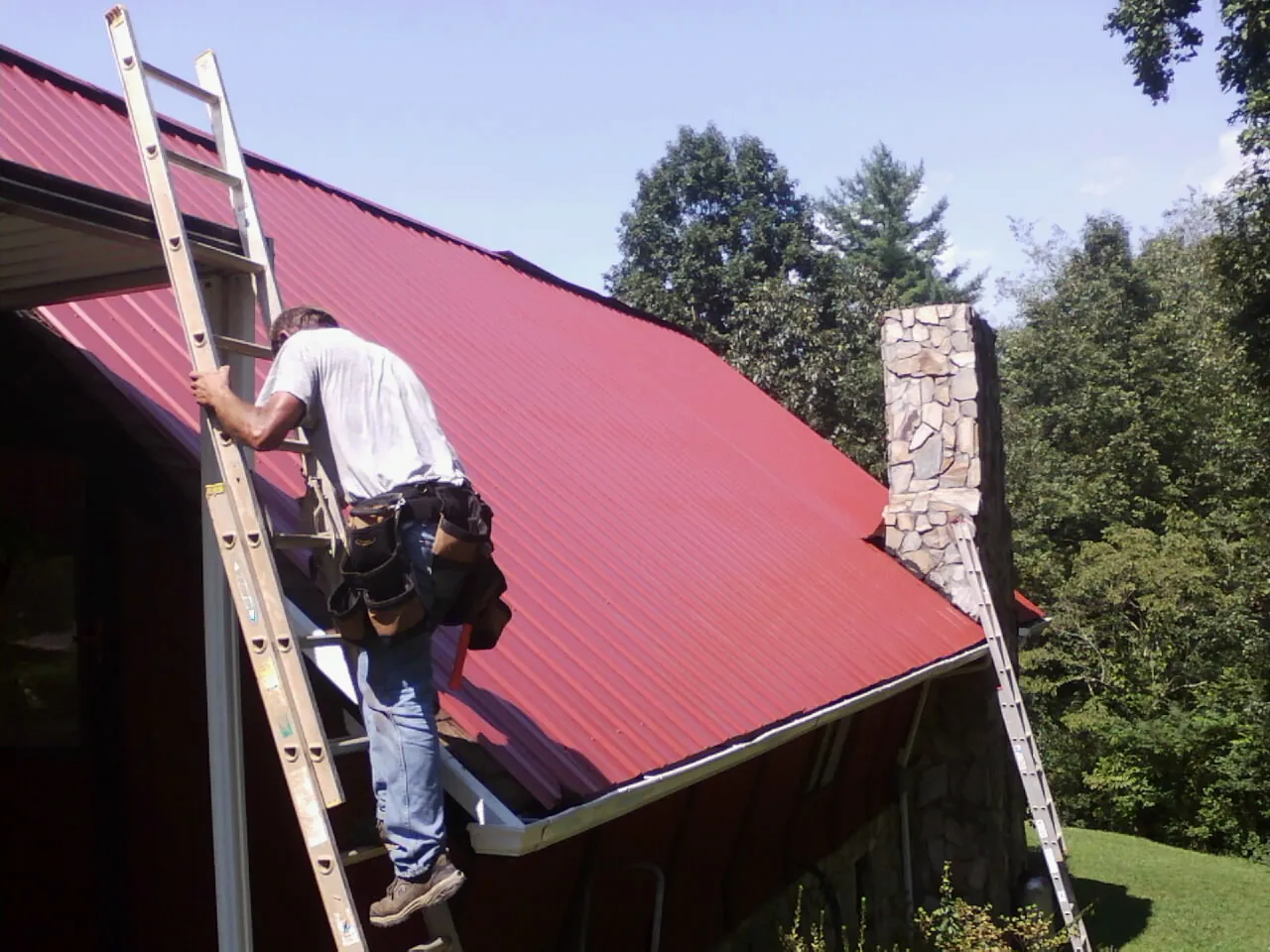 Roofer on a ladder beside a red metal roof and stone chimney — Soffit & Fascia Repair by Roofing Brothers in Broomes Island